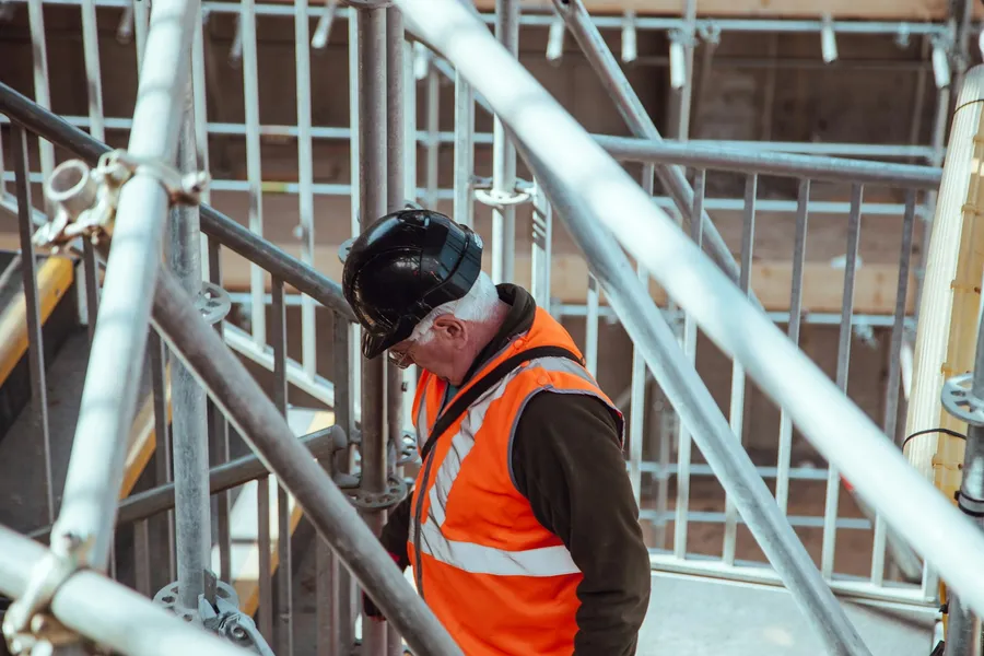 A construction worker in an orange hi-vis vest and hard hat on scaffolding
